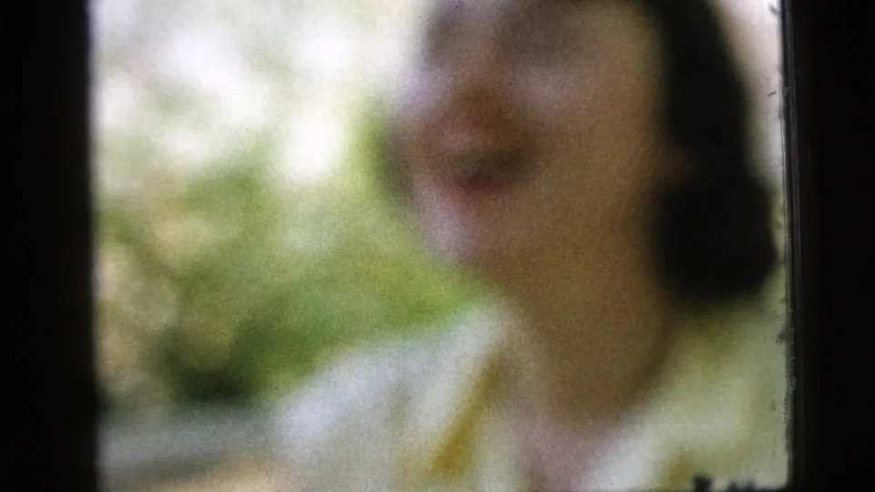 Soft-focus 1961 Camden home-movie frame of a woman smiling outdoors, partially obscured by blur and dark porch framing.