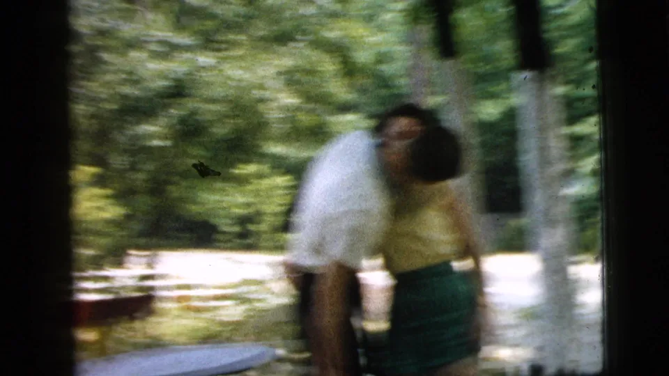 1961 Camden frame showing a man in a white shirt leaning toward a woman in green shorts near a white fence, red chair, and round table.