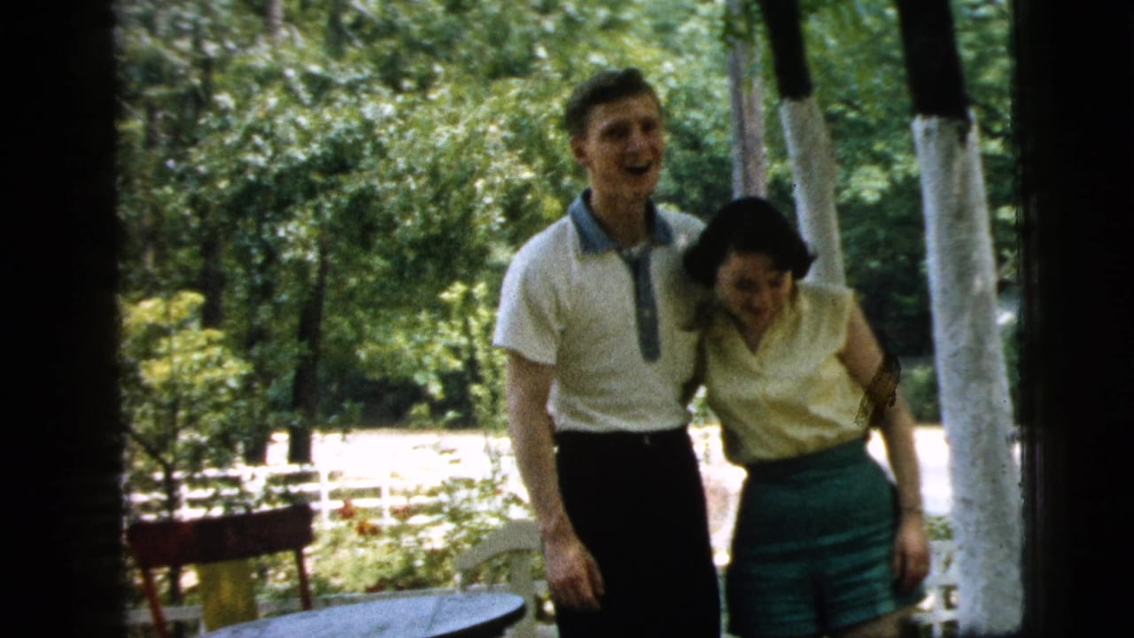 Young man stands with his arm around a woman on a shaded porch near trees and a small table.