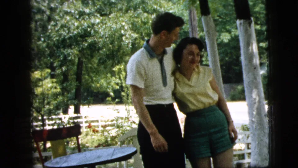 A young couple in casual summer clothing stands beside a small round table on a shaded porch in the 1961 Camden home movie.