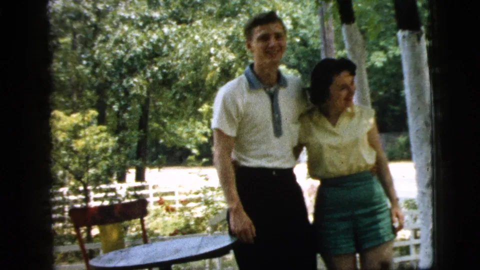The couple smiles together on the porch in Camden, New Jersey, framed by trees, a small table, and a white fence in the background.