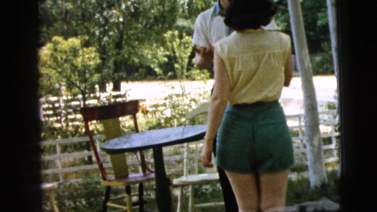 Woman in green shorts faces a man near a patio table and chairs in a leafy backyard.