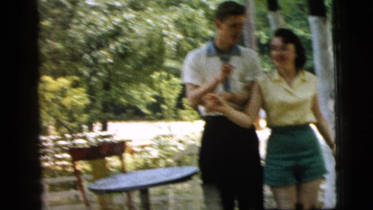 In the 1961 Camden backyard scene, the young couple smiles and holds hands beside a small round table and garden chairs.