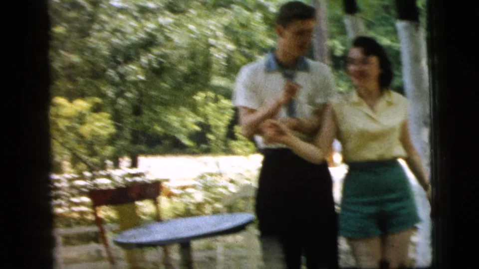 In the 1961 Camden backyard scene, the young couple smiles and holds hands beside a small round table and garden chairs.
