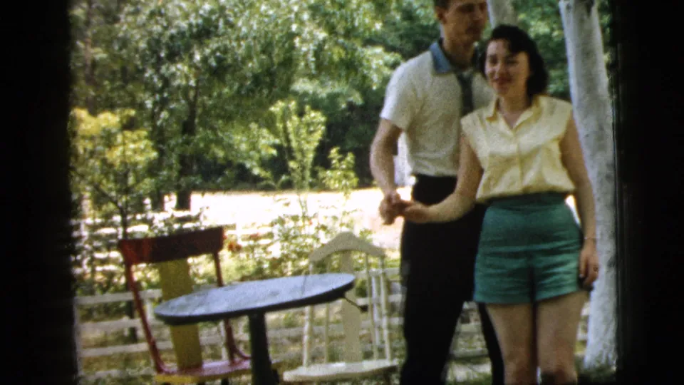 The 1961 Camden home movie shows the smiling couple holding hands in a leafy backyard with patio chairs and a white fence behind them.