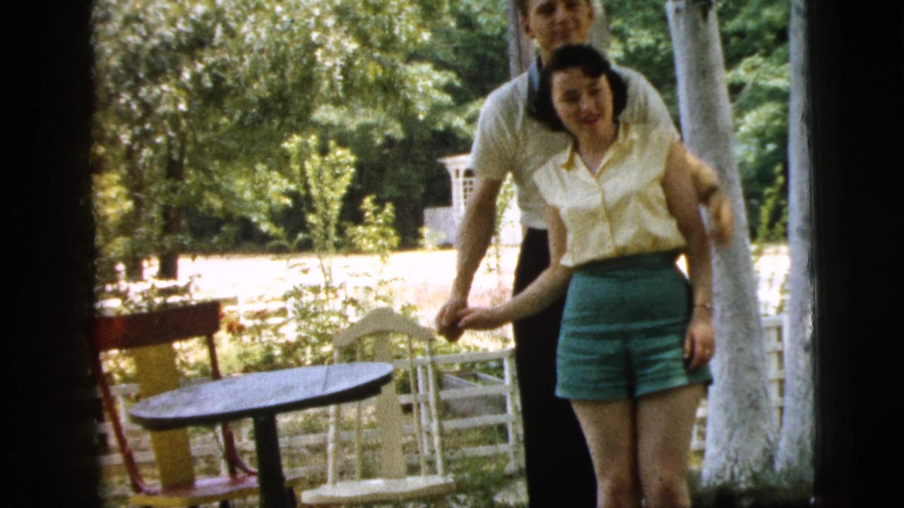 Young man stands behind a smiling woman near outdoor chairs and trees.