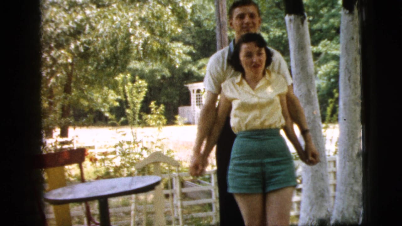Couple walks or poses together outside beside chairs and trees.