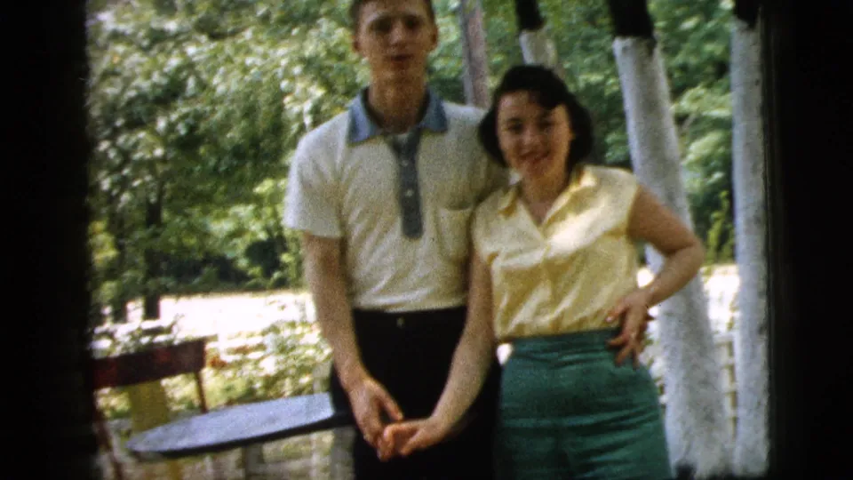 The woman smiles near the camera while holding the man's hand, with trees and patio furniture behind them.