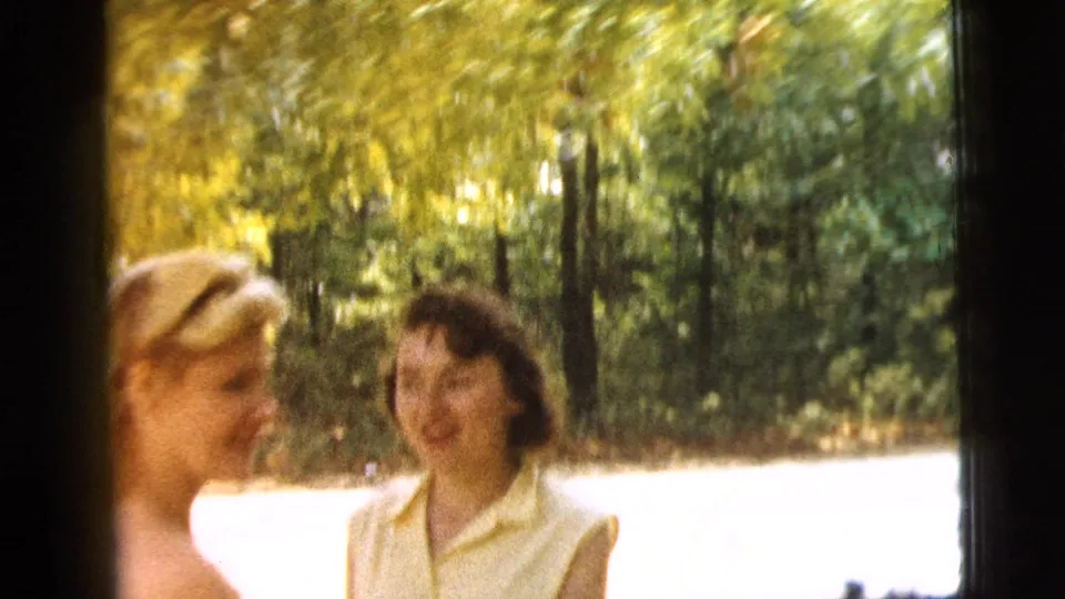 Soft 1961 Camden home-movie frame of the couple turned toward each other near trees and bright open ground.