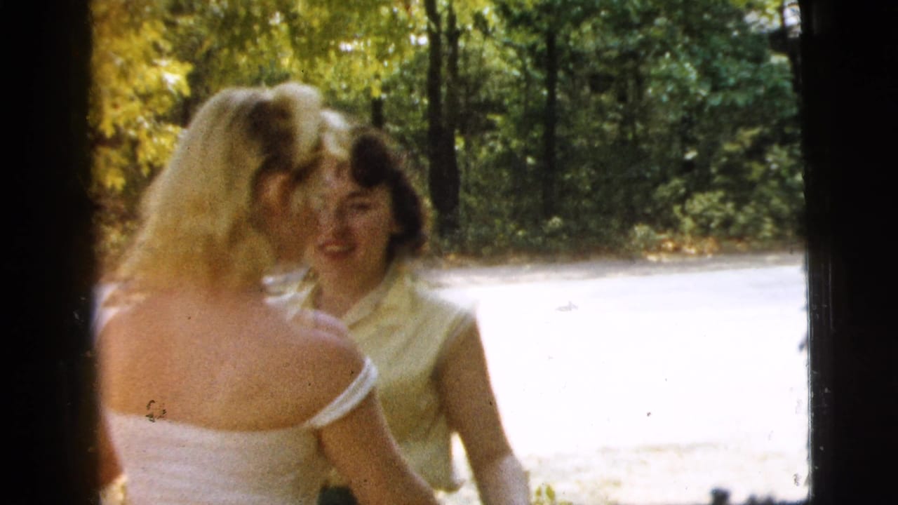 Two young women outdoors, one in a light dress and one in a pale top, near trees and a bright yard.