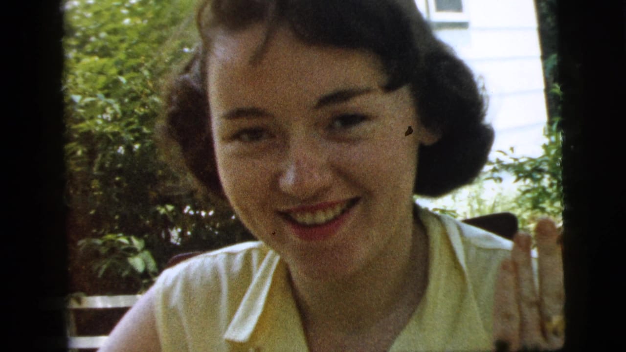 Close 1961 Camden home-movie view of a smiling young woman in a yellow sleeveless blouse, with shrubs and a white exterior wall behind her.