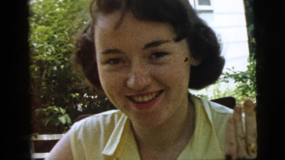 Close 1961 Camden home-movie view of a smiling young woman in a yellow sleeveless blouse, with shrubs and a white exterior wall behind her.