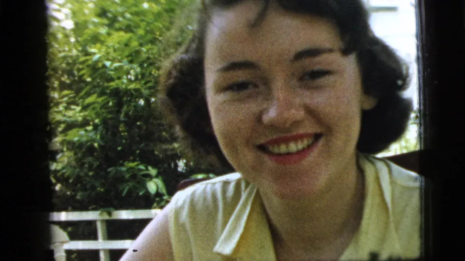 Close 8mm frame of a young woman smiling in a yellow blouse outdoors in Camden, with leafy shrubs and a white railing visible behind her.
