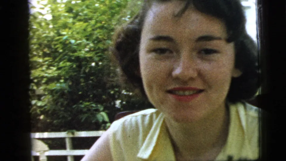 1961 Camden 8mm frame of a young woman facing the camera in a yellow sleeveless blouse, with greenery and a white railing in the background.