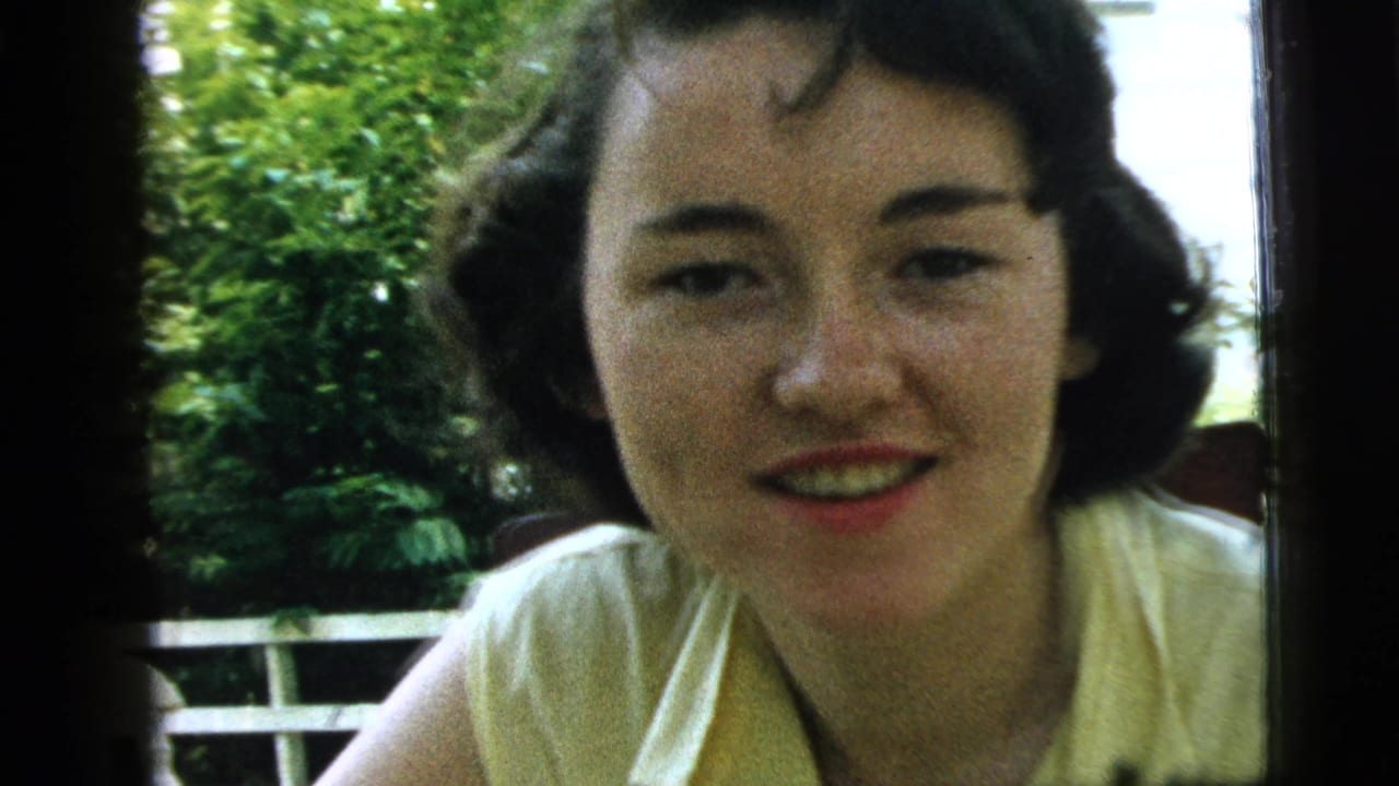 Young woman with dark curled hair smiles toward the camera beside a window and green foliage.