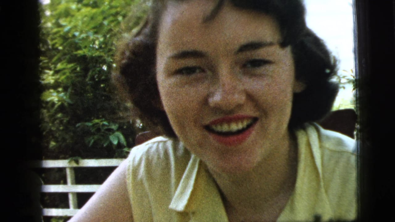 The 1961 Camden home-movie frame shows the woman smiling broadly, her face close to the camera against greenery and a porch rail.