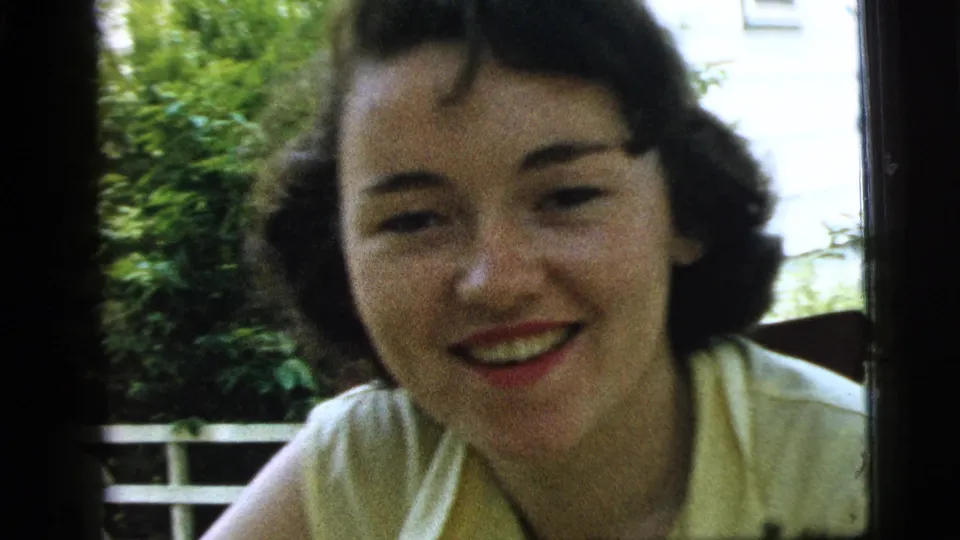 In this soft 8mm frame from Camden in 1961, the woman leans toward the camera while smiling near a window and outdoor plants.