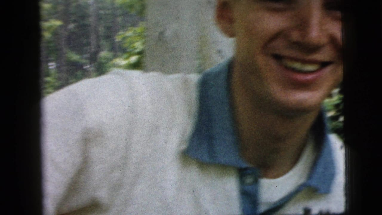 Smiling young man in a white shirt with blue collar outdoors near trees.