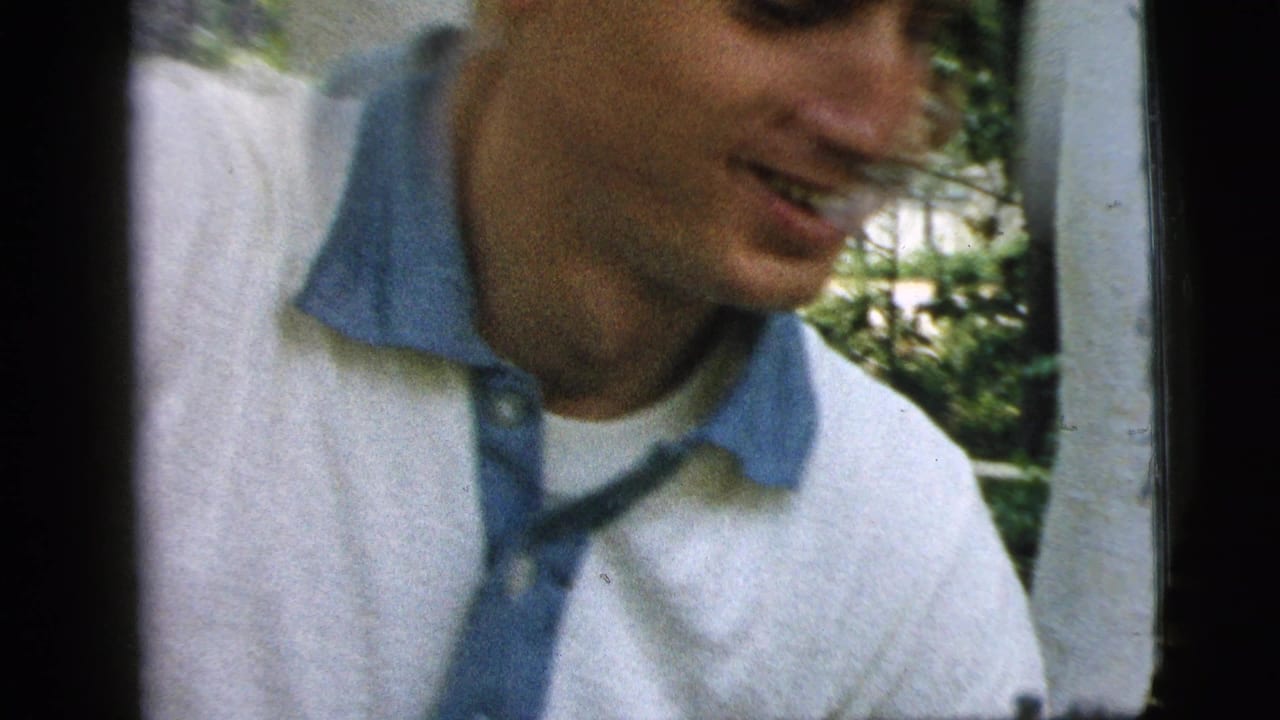 Young man in a white shirt with a blue collar leans toward the camera outdoors.