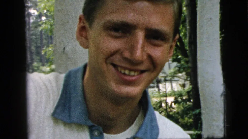 A smiling young man faces the camera in 1961 Camden footage, framed by a porch post and leafy background.