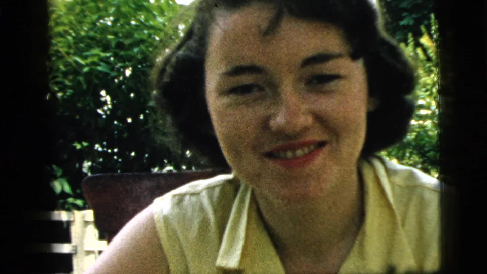 A young woman in a pale yellow sleeveless top smiles toward the camera in 1961 Camden home-movie footage, with greenery behind her.