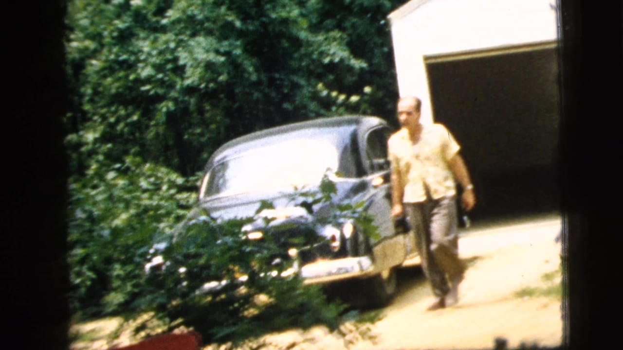 Man walking near a dark vintage sedan beside an open garage and leafy yard.