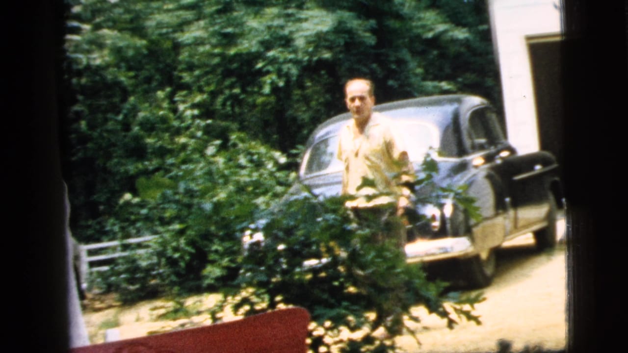 The man appears closer to the camera near a dark vintage car, with dense trees and shrubs framing the 1961 Camden yard scene.