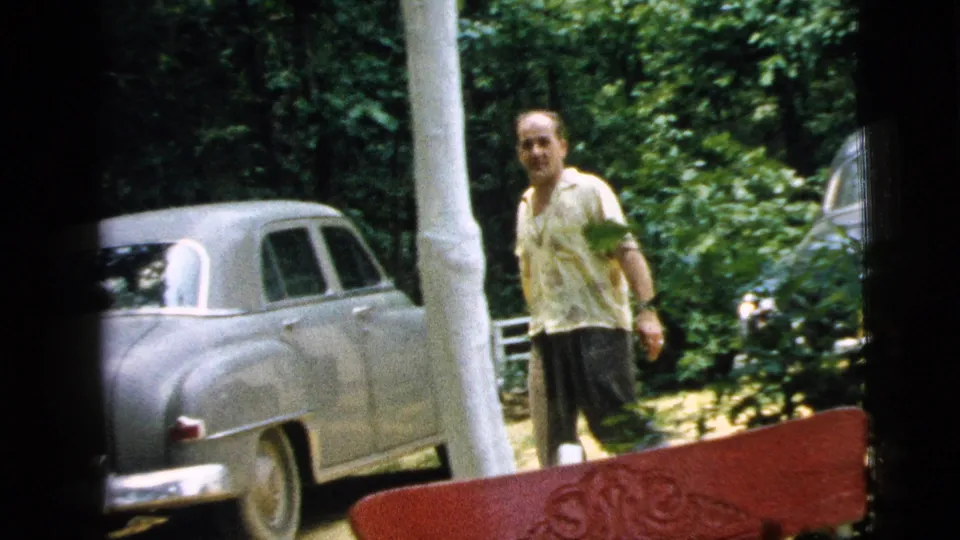 A man walks near white porch columns and a light-colored sedan, with a red outdoor chair edge visible in the 1961 Camden frame.