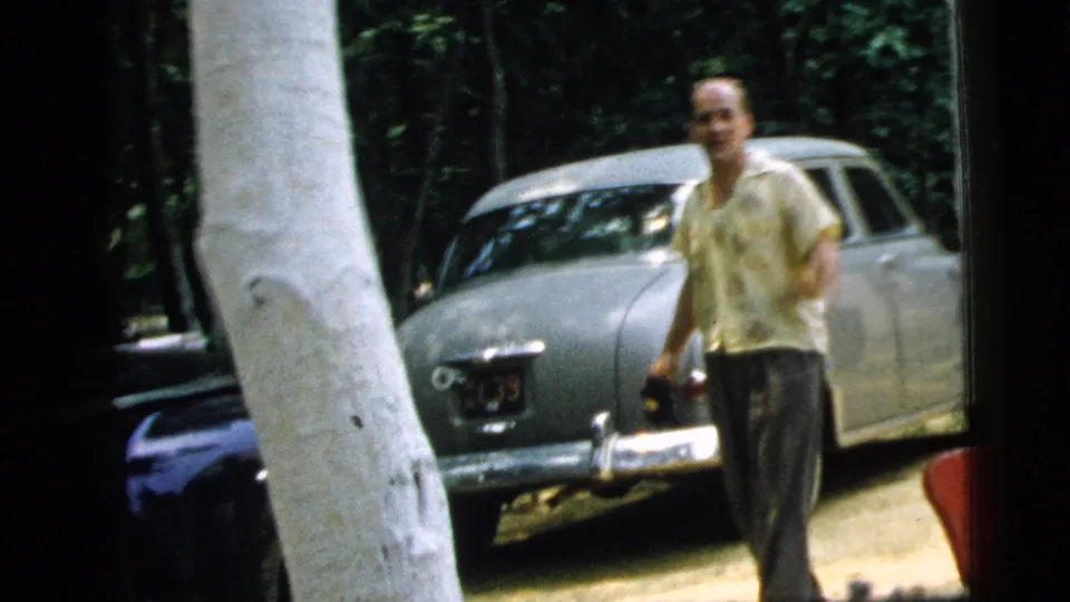 The man walks past the rear of a light-colored vintage sedan near white columns, with leafy trees in the Camden residential background.