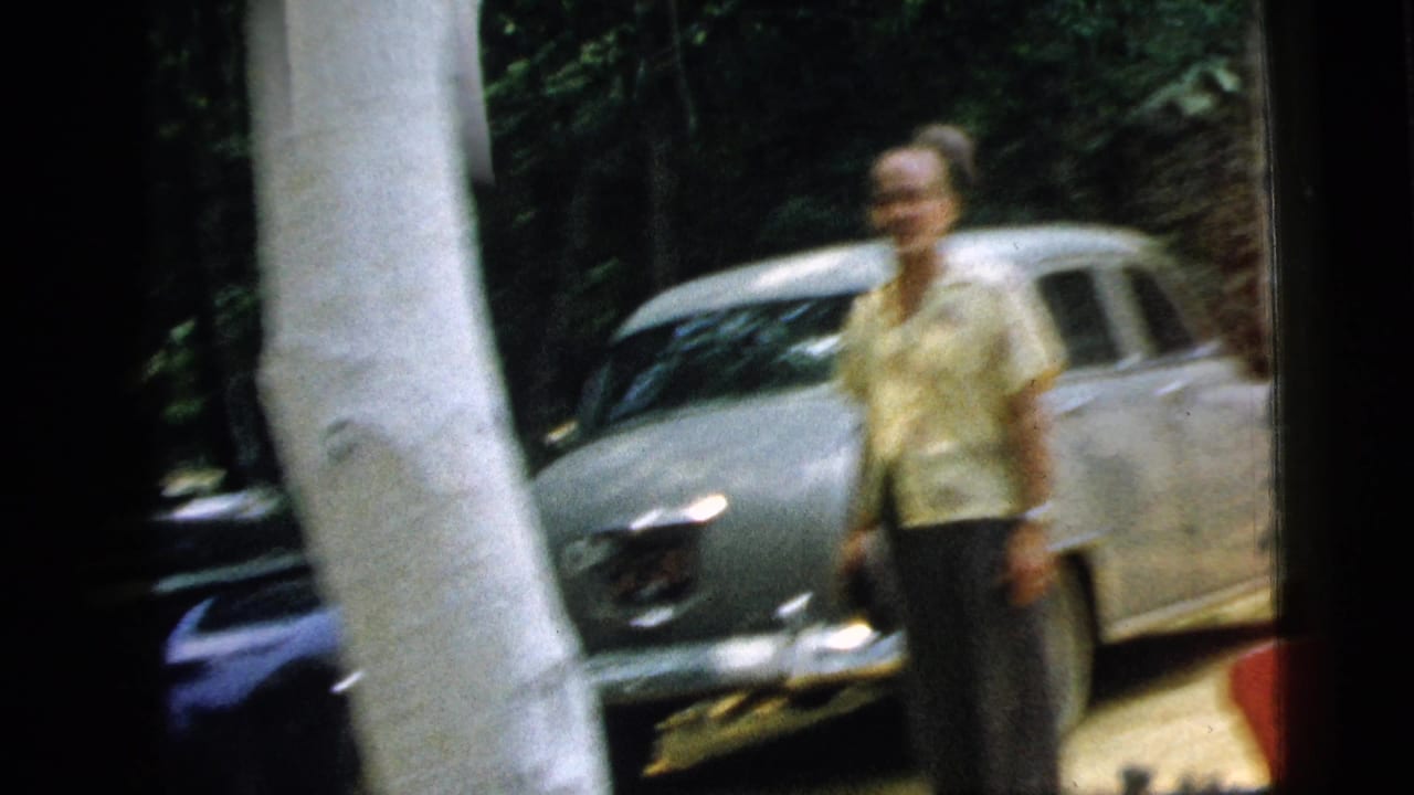 Man standing near a parked car outside a house, partly obscured by a porch post.