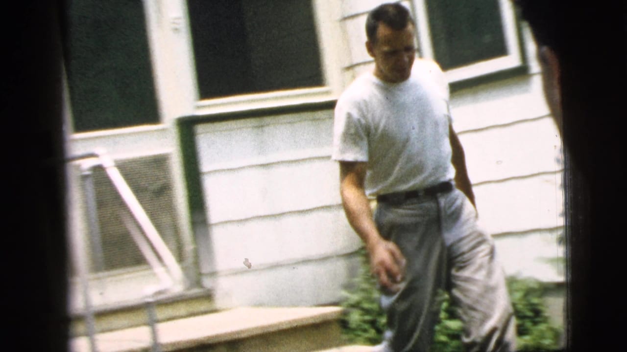 Adult man in a white shirt and light pants walks beside a Camden house exterior in 1961 home-movie footage.