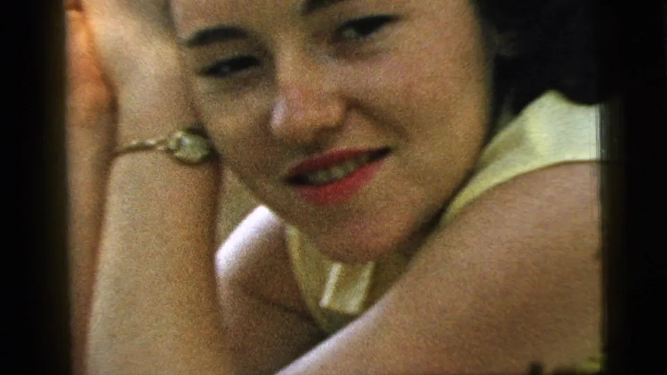 Adult woman in a yellow sleeveless top leans close to the camera, with her bracelet and smile visible in the 1961 Camden scene.