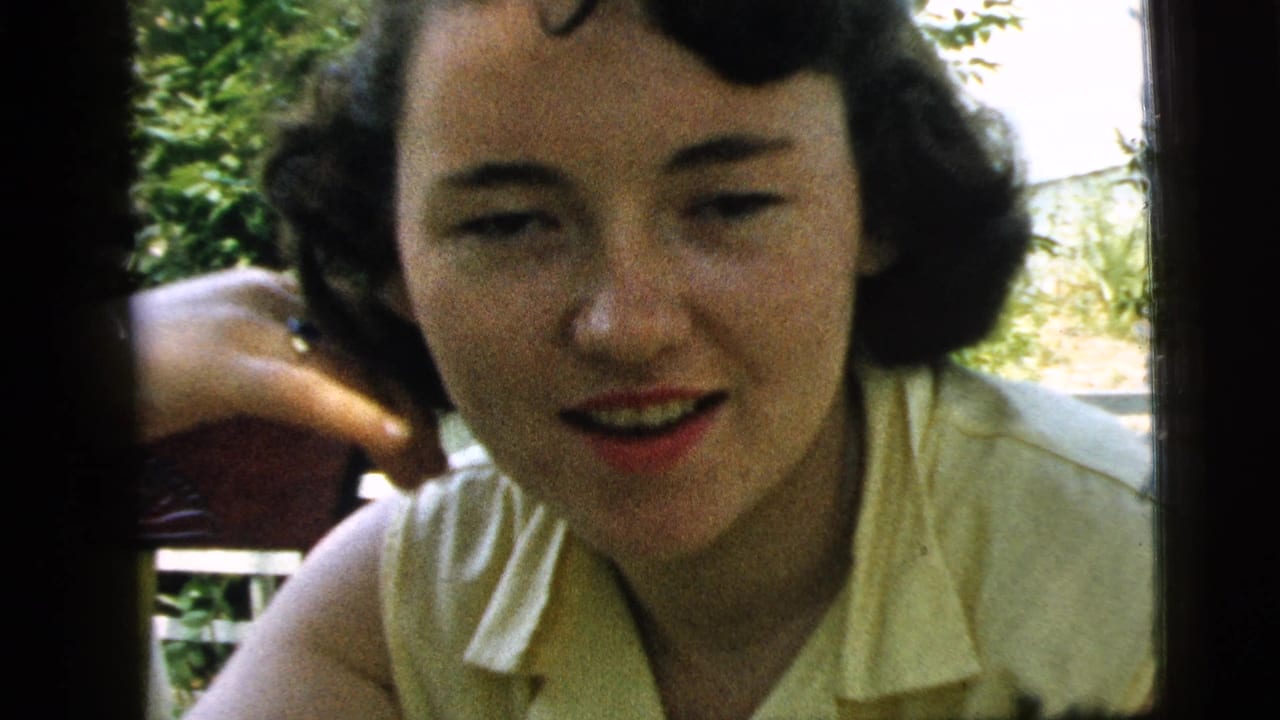 Young woman with dark curled hair looks toward the camera near greenery and a porch railing.
