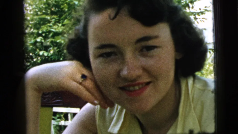 1961 Camden home-movie frame of a smiling young woman in a pale top, posed beside outdoor greenery.