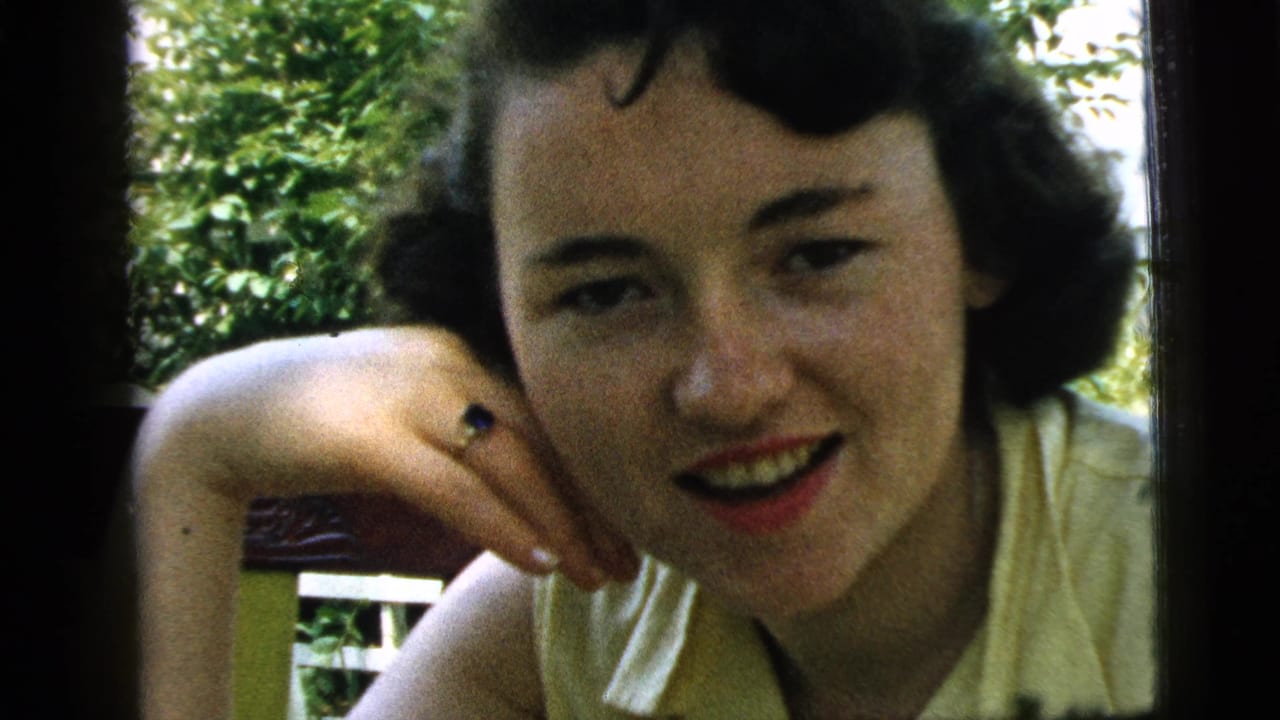 Young woman smiles with her hand near her face beside a window and greenery.