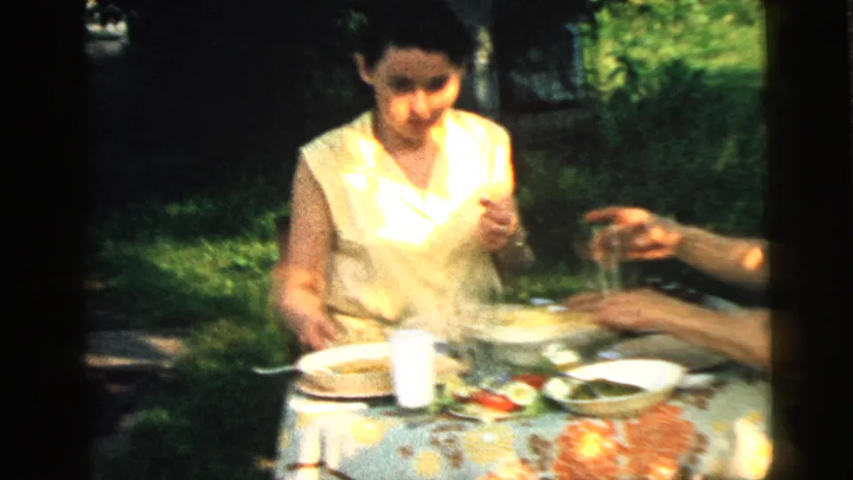 A woman sits at an outdoor lunch table in the 1961 Camden, New Jersey footage, with dishes and greenery visible around her.