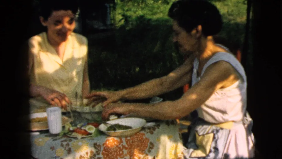 Two women sit at a floral-covered outdoor table in the 1961 Camden, New Jersey home movie, eating and handling dishes.