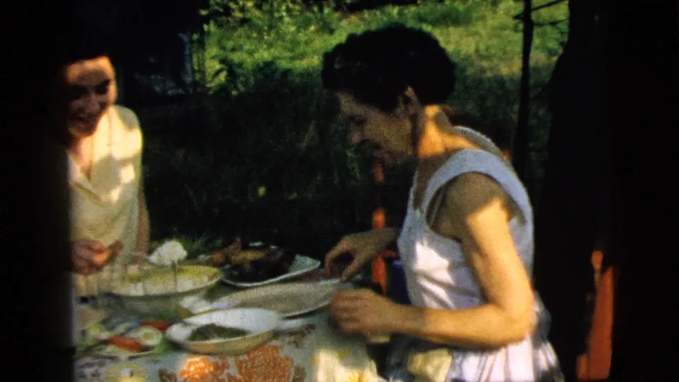 Two women continue an outdoor meal in the 1961 Camden, New Jersey footage, with plates, bowls, and shaded greenery behind them.