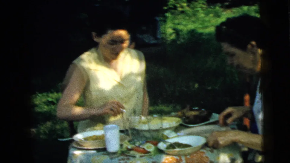 In 1961 Camden, New Jersey home-movie footage, two women sit at a garden table with lunch plates, glasses, and a floral tablecloth.