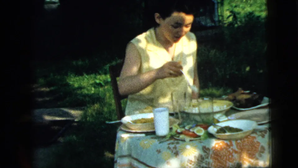 A woman in a light sleeveless dress eats at an outdoor lunch table in Camden, New Jersey, with plates, glasses, and garden greenery visible.