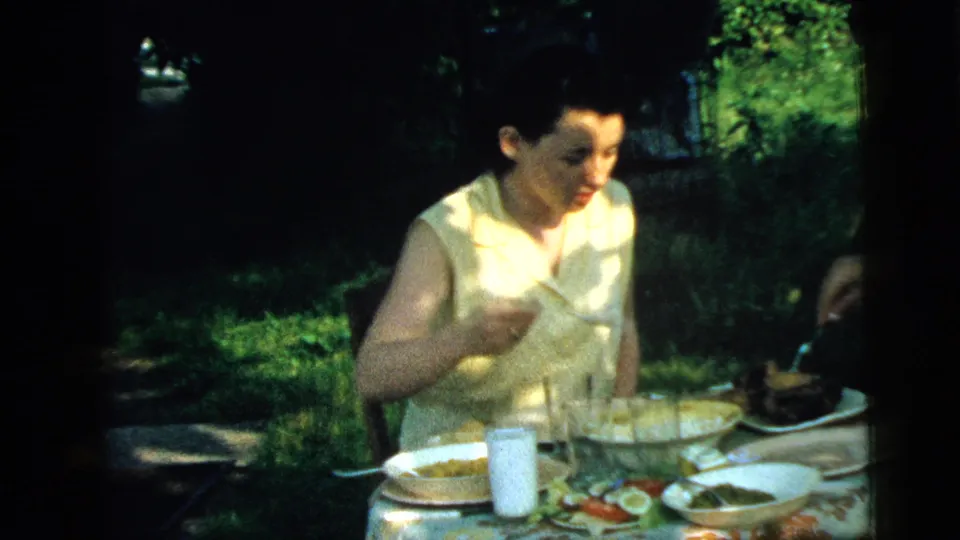 A woman looks toward the lunch table in 1961 home-movie footage, with plates, glasses, a white cup, and a shaded garden behind her.
