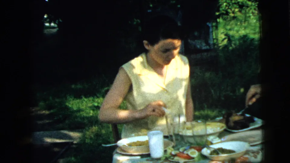 A woman eats at a shaded outdoor lunch table while another person is partly visible at the edge of the 1961 Camden home-movie frame.