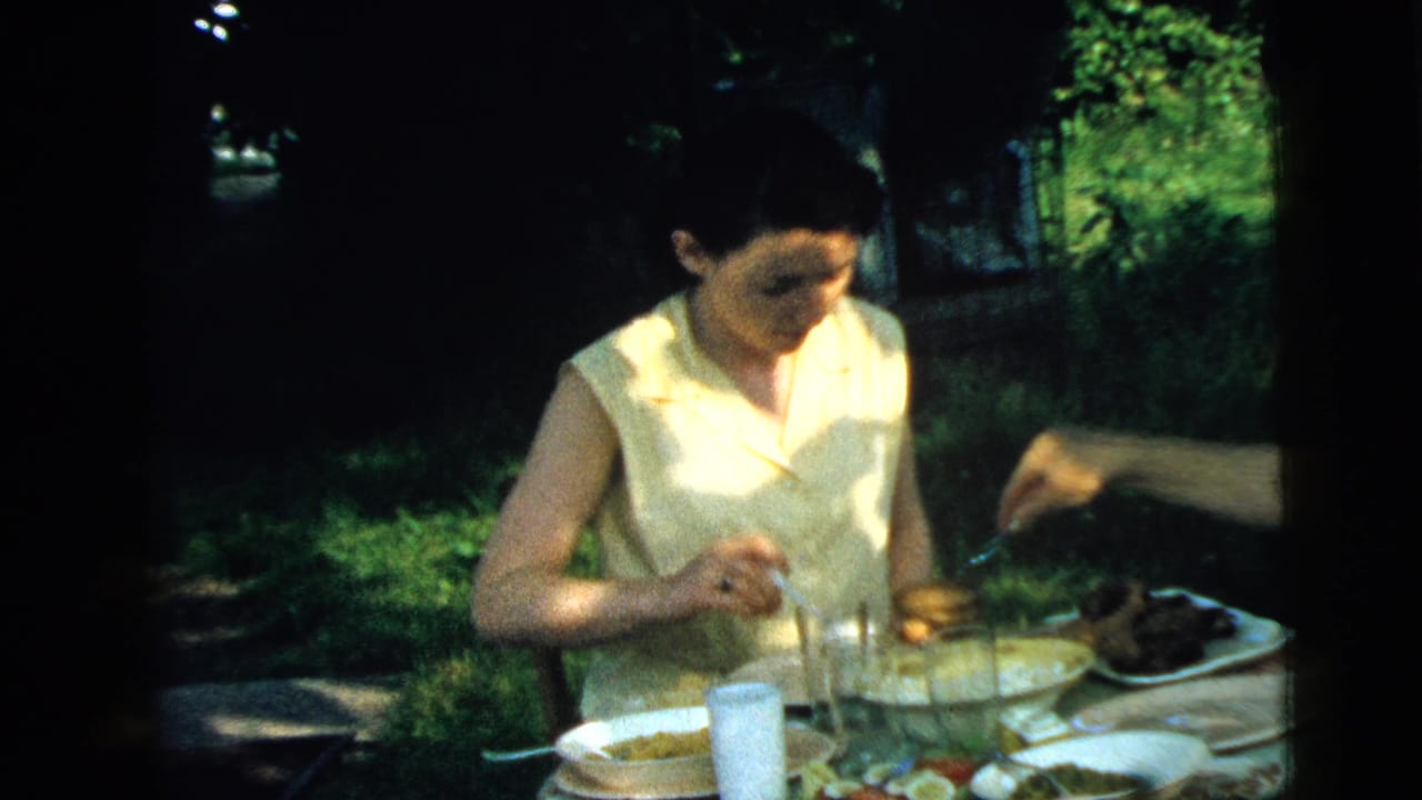 Woman in a pale sleeveless dress eating at an outdoor table in a grassy yard.