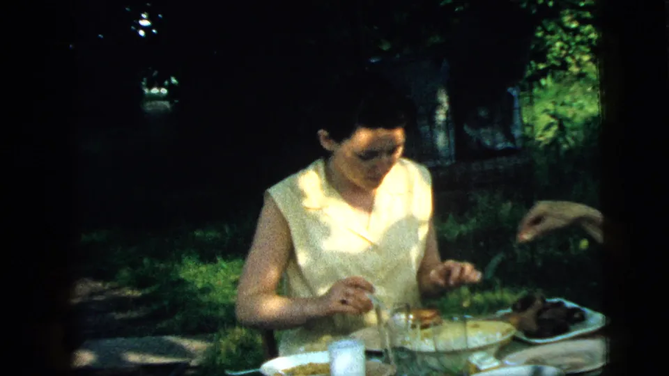 1961 Camden backyard meal frame showing a seated woman, plates, glasses, and an arm reaching across the table.