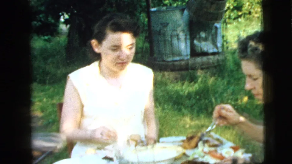 1961 Camden home-movie frame of two women sharing an outdoor meal at a table in a grassy yard.
