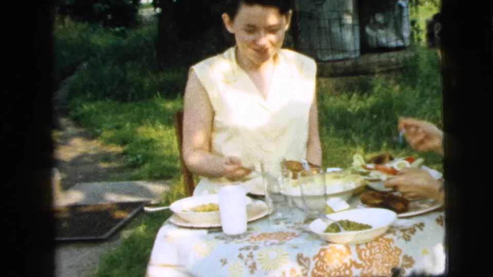 A woman in a pale sleeveless top looks down at food on a garden lunch table, with dishes, a glass, and greenery visible in the 1961 home movie frame.