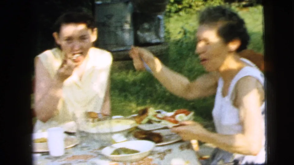 Two women appear to laugh and eat at a garden lunch table in 1961 Camden, New Jersey, surrounded by plates, serving dishes, grass, and filtered sunlight.
