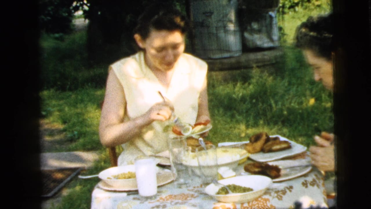 A woman in a pale sleeveless dress eats at an outdoor table while another person sits nearby.