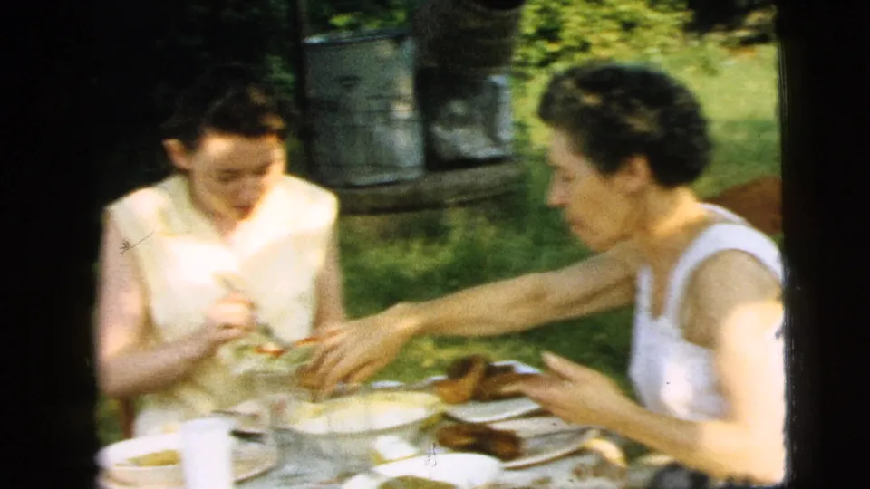 Two adult women at a Camden garden table in 1961 serve and share food during a casual outdoor meal.