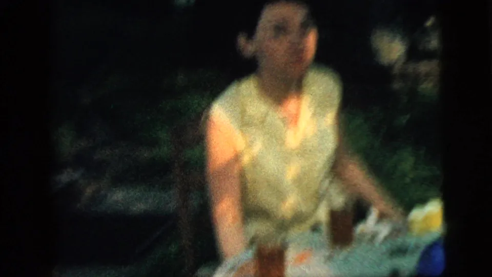 In supported 1961 Camden home-movie footage, a brown-haired woman sits outdoors in a yellow sleeveless blouse near glasses on a table.